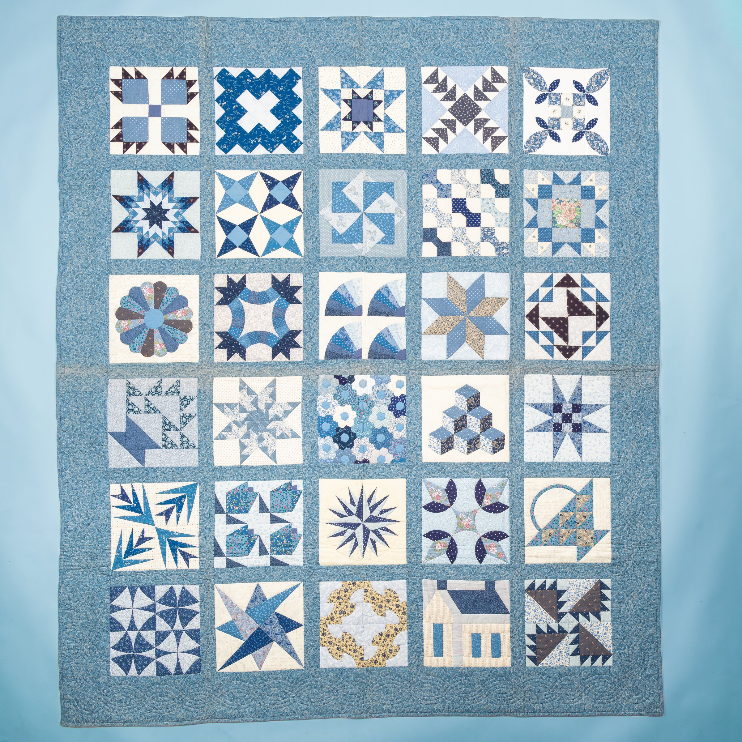 A blue sampler quilt with many geometric quilt blocks on a blue studio backdrop.